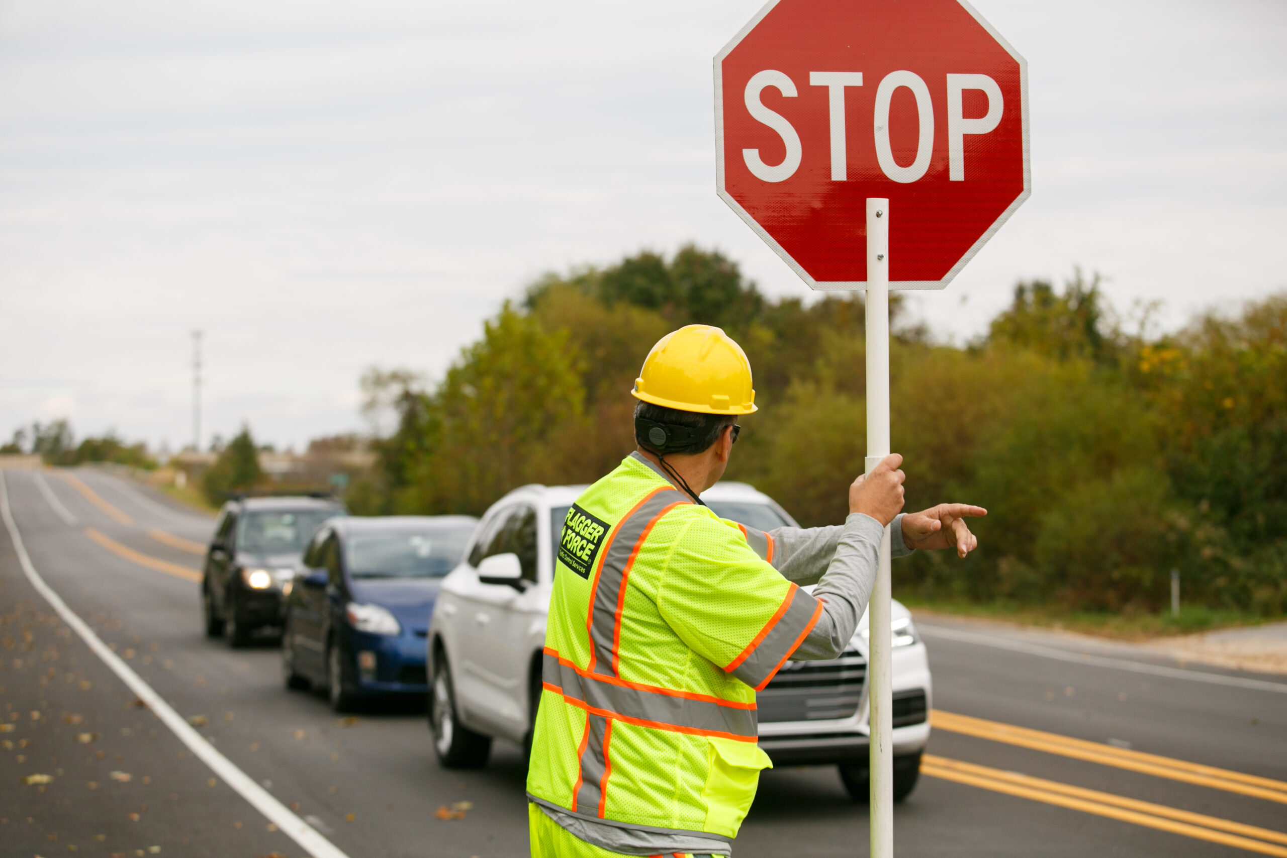 A Flagger Force employee working outdoors, wearing a hard hat, a reflective vest, holding a sign, and directing traffic.